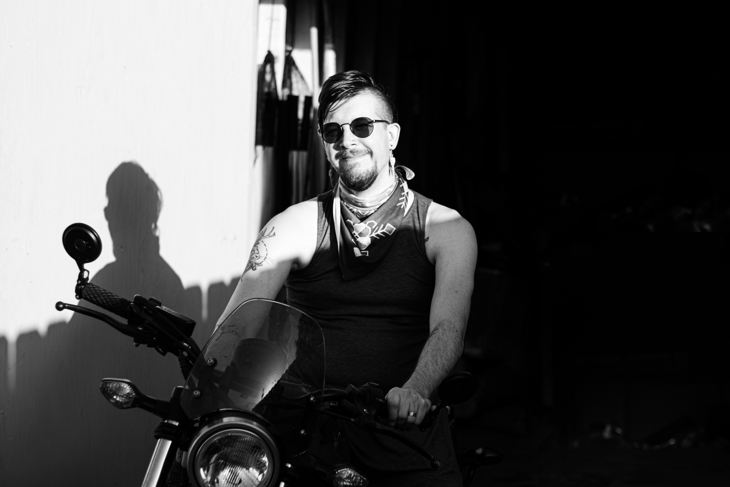 Black and white picture of Hilary wearing a sleeveless top, round dark glasses, dangly earrings, and a bandana. They have a short goatee and an undercut hairstyle, and they're smiling. They are sitting astride a motorcycle with a round headlight and a small windshield. They are in a ray of sunlight that cuts across their shoulders, casting a shadow on the white background to their left, while leaving the remaining two thirds of the backdrop dark and featureless.
