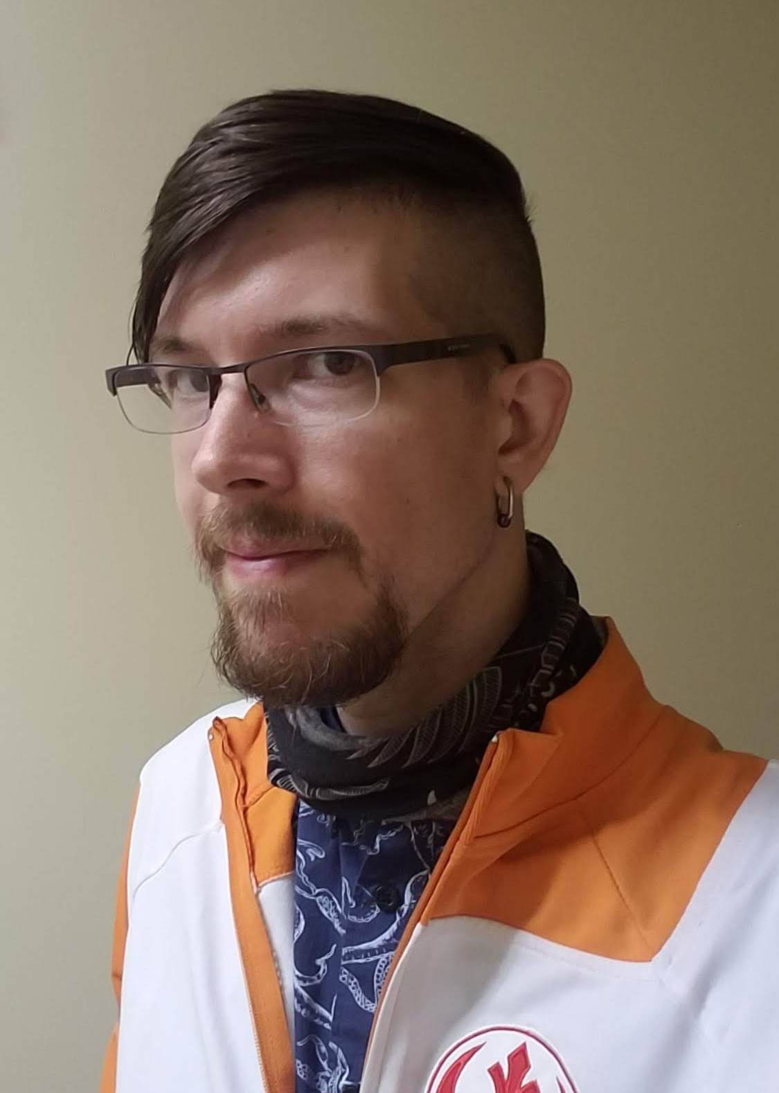 Image of the host wearing half-framed rectangular glasses, a blue shirt, and an orange Rebel Alliance jacket.