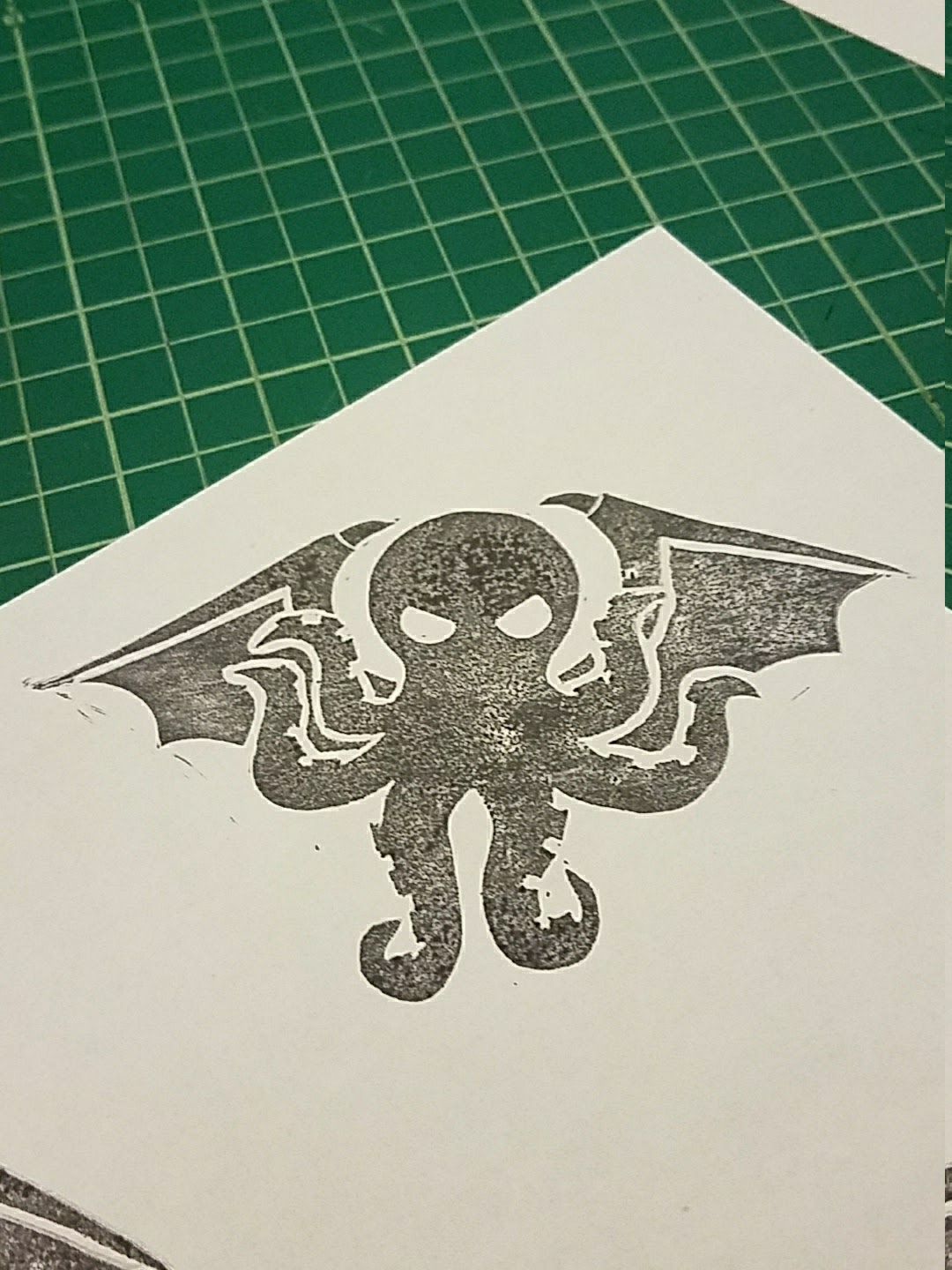 A block-printed image of Cthulhu's head and wings in a mottled black on a corner of a piece of white paper. The paper is resting on a gridded green cutting mat.