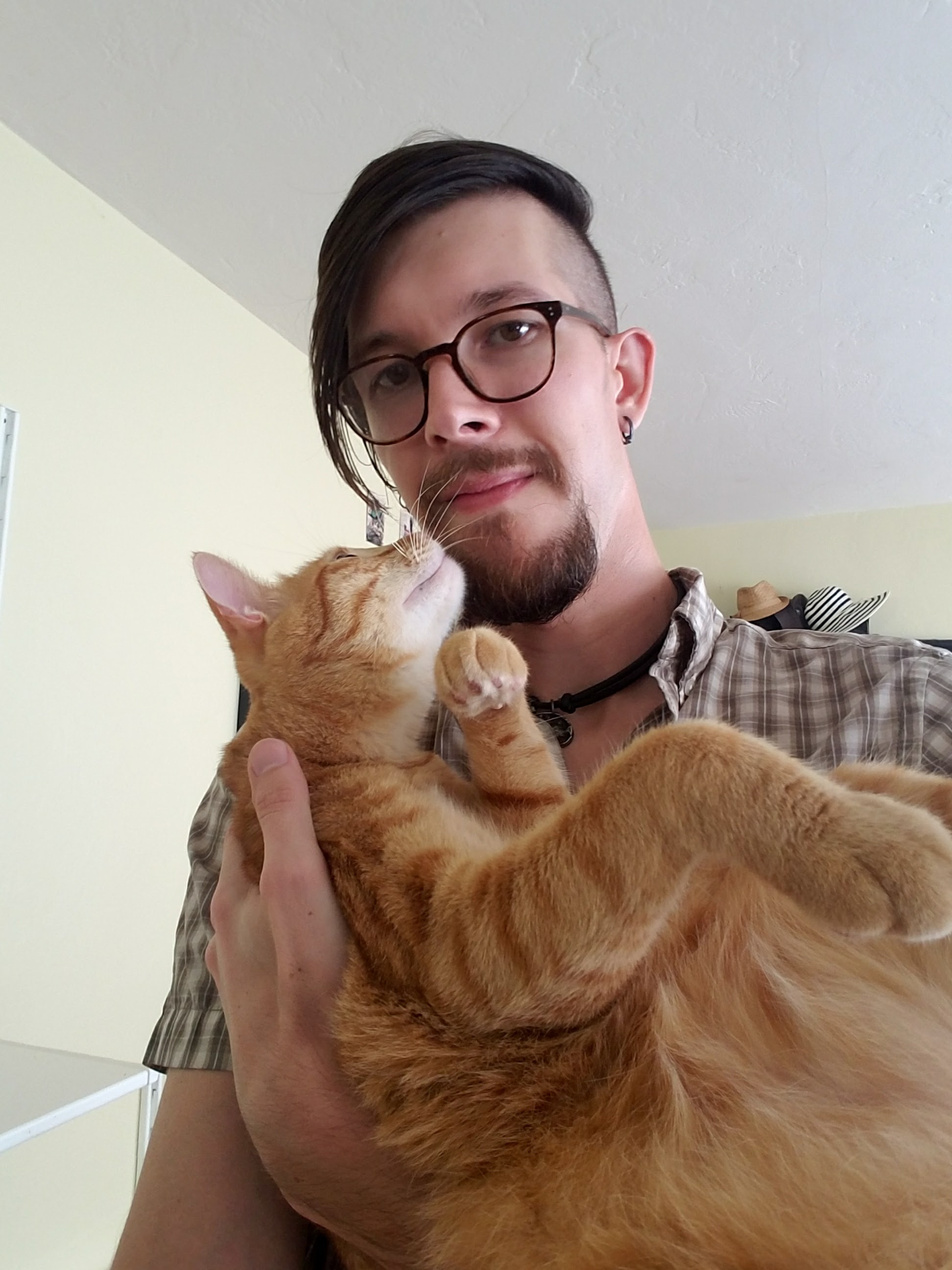 Image of Hilary, wearing round, brown glasses, their hair styled in an undercut and swept to the right of their face, holding an orange cat.